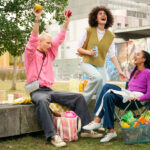 Drei junge Menschen sitzen lachend bei einem Picknick im Grünen vor einem modernen Gebäude. Einer hält Obst in die Luft, während neben ihnen Einkaufstaschen, Lebensmittel und ein Fahrrad stehen